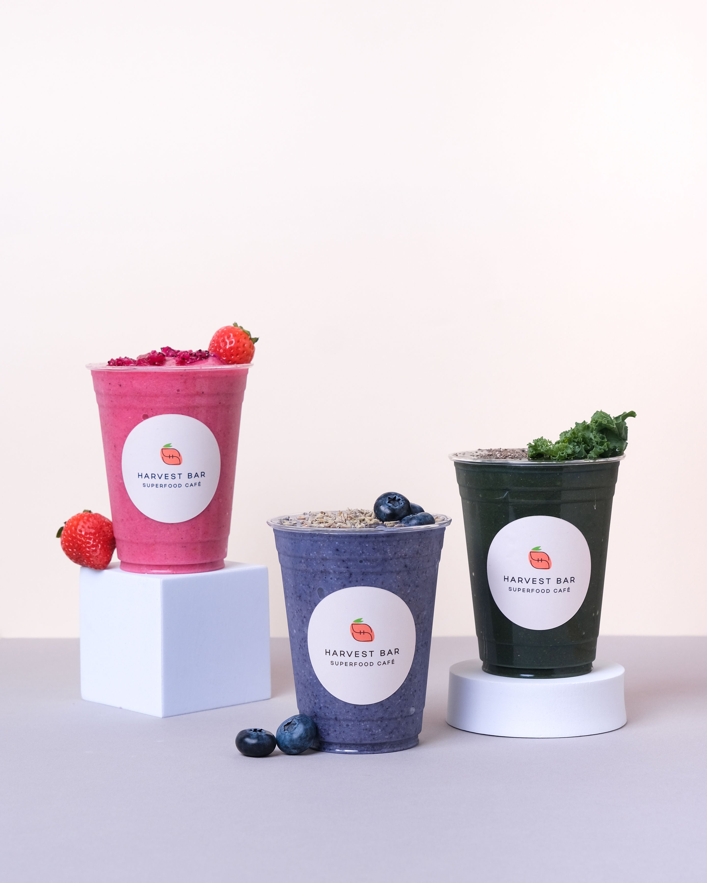 Three smoothie cups from Harvest Bar on minimalist white pedestals against a light beige background. The left cup is pink with strawberries, the middle is blue-purple with blueberries, and the right is dark green with kale, all with matching branded labels.